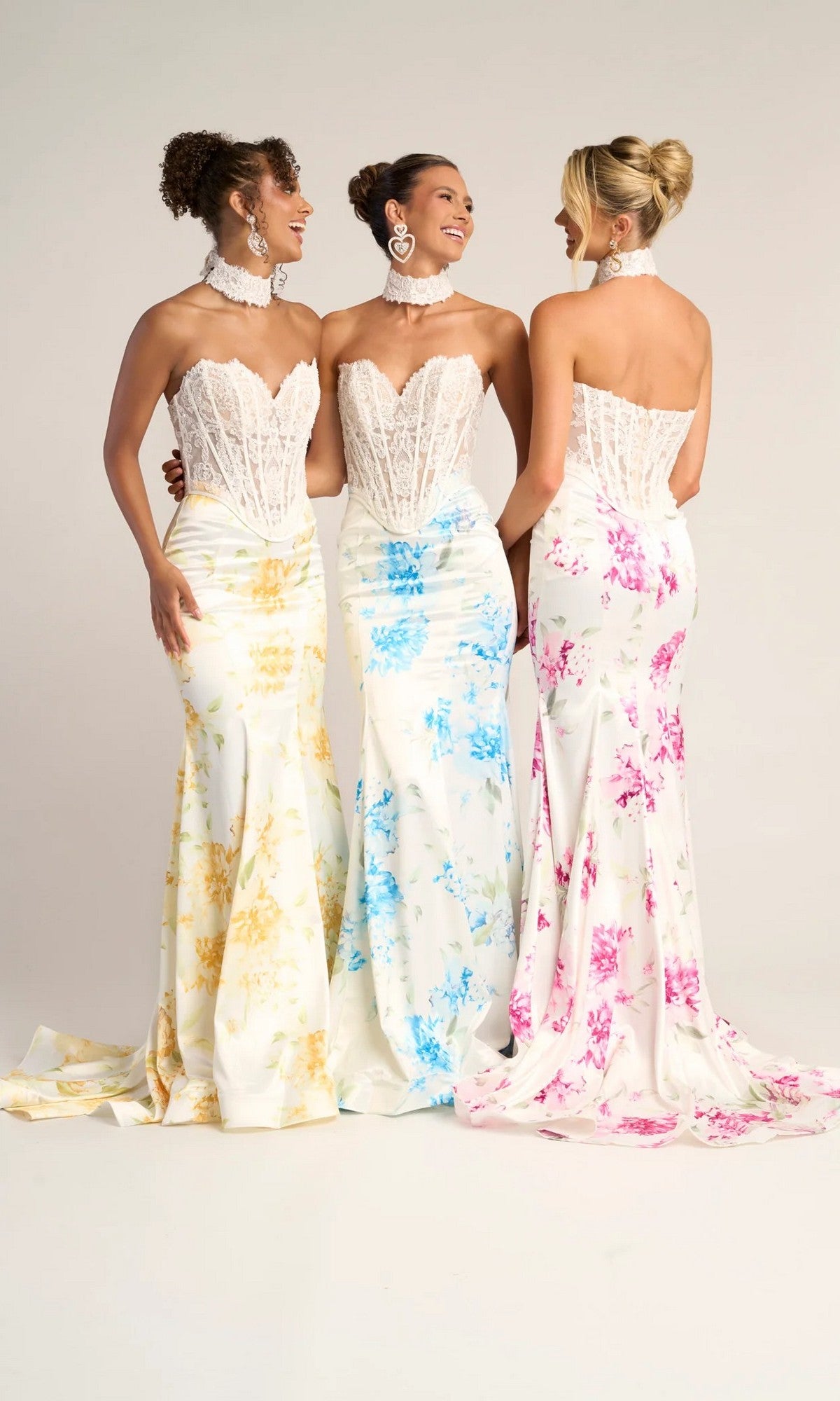 Three women stand together in Portia and Scarlett PS26299 long formal dresses, featuring white strapless lace corset gowns with yellow, blue, and pink floral patterns. They are smiling at each other against a plain background.